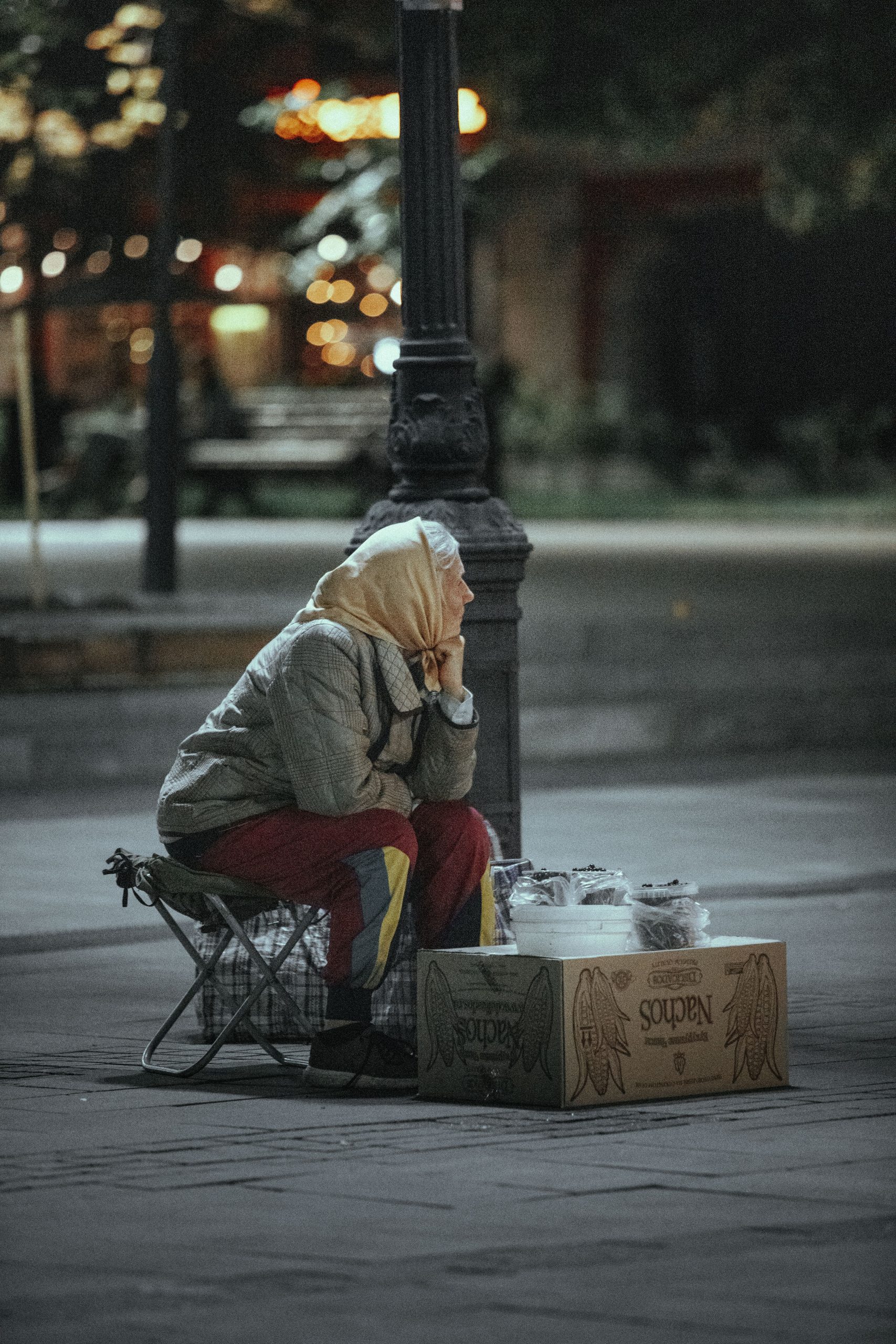 An elderly woman sitting at nighttime, selling items on a street corner.